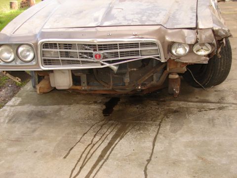1973 Ford Ranchero - Front-end damage on a faded tan 1970s Ford with quad round headlights and an eggcrate grille wearing a red Ford crest emblem; the bumper is…