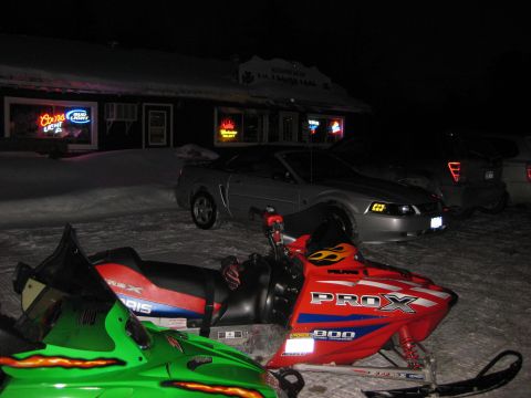 1995 Ford Bronco - Two snowmobiles are parked in front of a building at night. A silver Ford Mustang is parked behind the snowmobiles.