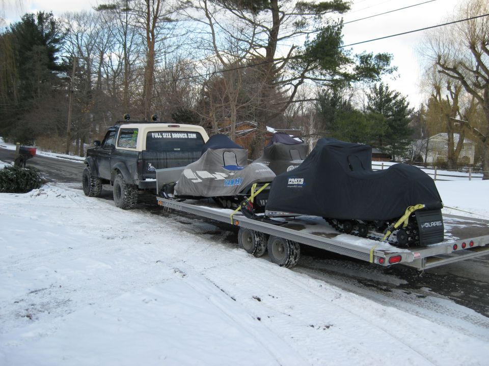 Lifted Ford Bronco towing a trailer with two snowmobiles in the snow.