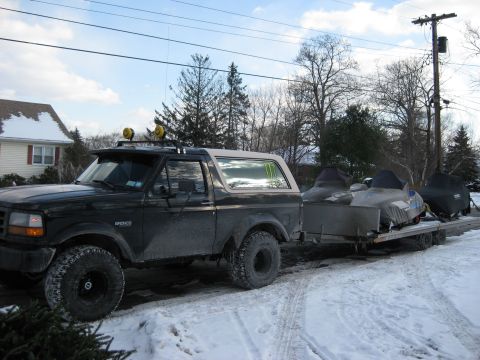 1995 Ford Bronco - A black Ford Bronco is parked on a snowy road, towing a trailer with three snowmobiles covered by tarps. The vehicle has two yellow lights…