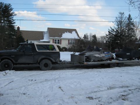 1995 Ford Bronco - A black Ford Bronco is parked on a snow-covered road, towing a trailer with two snowmobiles covered by tarps. The Bronco has a tan top and…