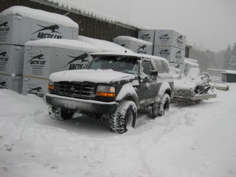 1995 Ford Bronco - A dark-colored Ford Bronco, covered in snow, is parked outside a building with stacks of Arctic Cat snowmobile boxes. A snowmobile trailer…