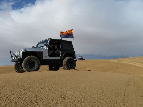 2004 Jeep Wrangler - A silver Jeep Wrangler Rubicon with a black soft top is parked on a sand dune, flying an Arizona state flag. Another off-road vehicle is vi…
