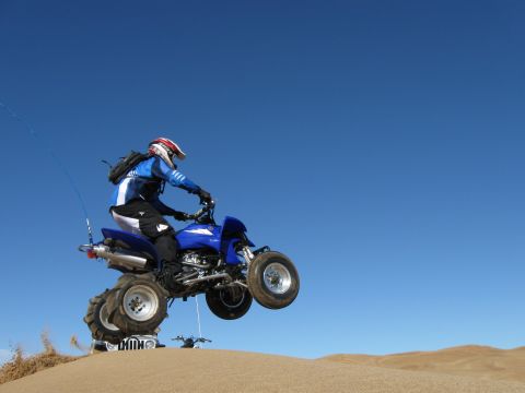 2004 Jeep Wrangler - A person wearing a blue and white racing suit and a red and white helmet rides a blue ATV through the air over sand dunes under a clear blu…