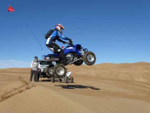 2004 Jeep Wrangler - A person in a blue and white racing suit on a blue ATV is mid-air over sand dunes. Another person stands next to a Jeep Wrangler in the bac…