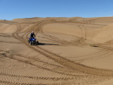 2004 Jeep Wrangler - A person rides a blue ATV through sand dunes under a clear blue sky. Tire tracks are visible in the sand, indicating previous activity.