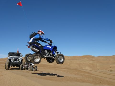 2004 Jeep Wrangler - A blue ATV is captured mid-air as it jumps over a smaller ATV, with a white Jeep Wrangler parked behind them in the sand dunes.