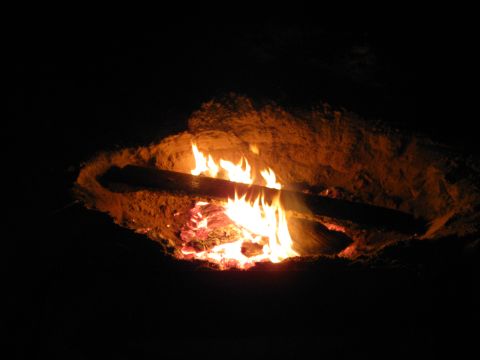 2004 Jeep Wrangler - A campfire burns brightly at night, with flames illuminating the surrounding sand and logs.