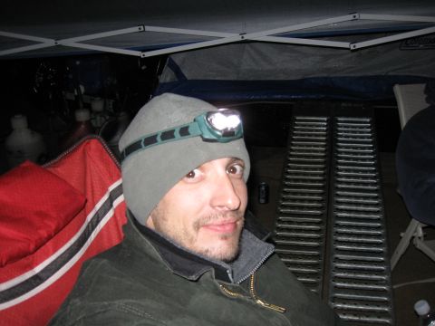 2004 Jeep Wrangler - A person wearing a headlamp and a jacket is sitting in front of a tent. There are some red and white striped fabric and metal ramps visible…