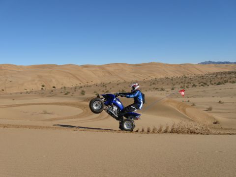 2004 Jeep Wrangler - A person riding a blue ATV performs a wheelie on a sand dune, kicking up sand. The ATV has a red flag attached to a whip antenna.