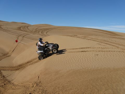 2004 Jeep Wrangler - A person rides a white and black ATV up a sand dune, with a red flag attached to the back. Tire tracks are visible on the sand dunes under…