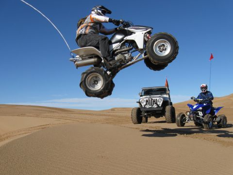 2004 Jeep Wrangler - A person on a white quad bike jumps over a sand dune, with a silver Jeep Wrangler and a blue quad bike on the sand below.