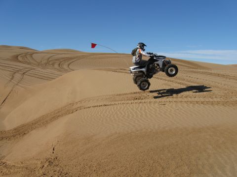 2004 Jeep Wrangler - A person on a white ATV is airborne over sand dunes, with a red flag attached to the vehicle. The ATV is mid-jump with its rear wheels off…