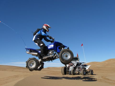 2004 Jeep Wrangler - A person on a blue ATV is mid-air over sand dunes, performing a jump. Another ATV and a Jeep Wrangler are visible in the background.
