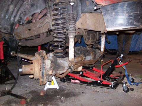 1986 Ford Bronco - The front axle and suspension components of a vehicle are supported by jack stands and a red hydraulic lift. The vehicle appears to be unde…