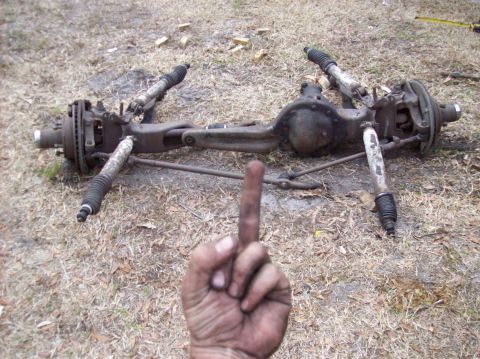 1986 Ford Bronco - A disassembled front axle assembly with shocks and brake rotors is laid out on the ground. A dirty hand is in the foreground giving the mid…