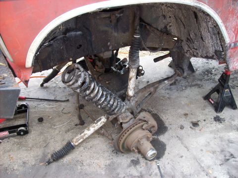 1986 Ford Bronco - The front suspension of a red vehicle is disassembled, with the coil spring and shock absorber lying on the ground next to the vehicle fram…