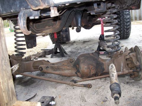 1986 Ford Bronco - The underside of a vehicle is shown with the front axle assembly removed and supported by jack stands. Suspension components like springs a…