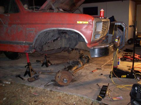 1986 Ford Bronco - A red Ford truck is jacked up with its front axle and suspension components removed and lying on the ground. Tools and a floor jack are sca…
