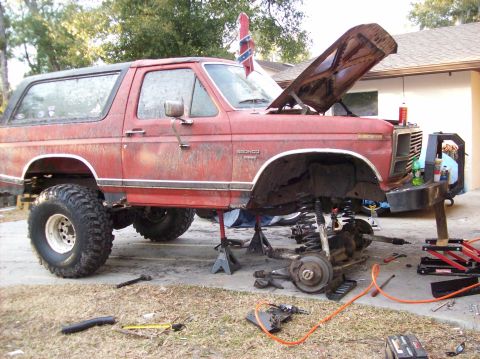 1986 Ford Bronco - A red 1980s Ford Bronco is undergoing repairs with its hood open and front end disassembled. The vehicle is elevated on jack stands, reveal…
