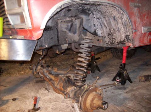 1986 Ford Bronco - The front suspension of a red 1986 Ford Bronco is disassembled, with the axle and coil spring removed and supported by jack stands. Tools l…