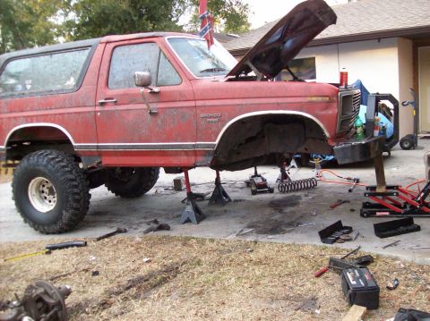1986 Ford Bronco - A red Ford Bronco is jacked up and appears to be undergoing maintenance or modification. The hood is open, and various tools and equipment…