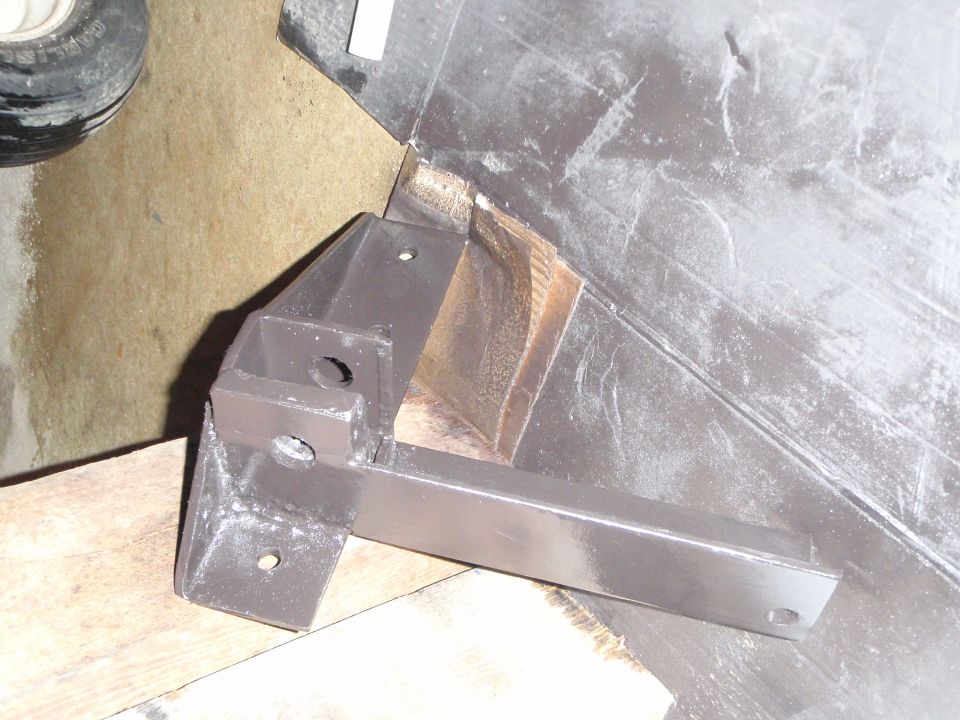 Dark gray metal bracket with holes mounted on wood, with tire and concrete floor in background.