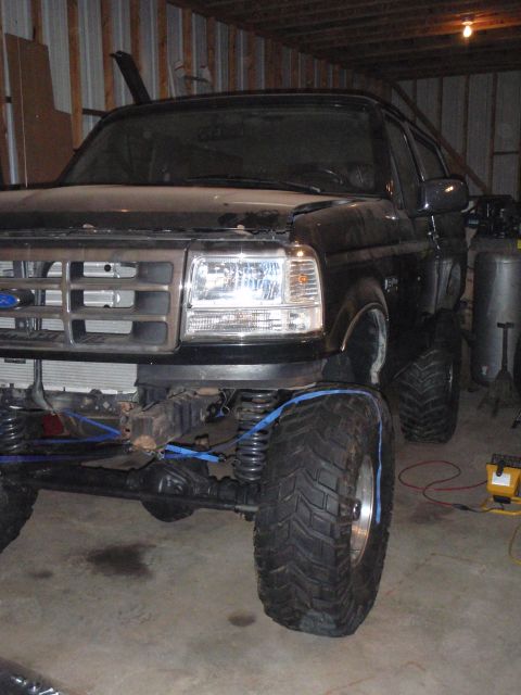 1996 Ford Bronco - A dark-colored Ford Bronco is parked inside a garage, showing its front end and lifted suspension with large off-road tires secured by blue…