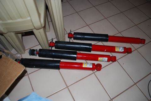 2008 Jeep Wrangler - Set of four red and black aftermarket shock absorbers laid out on a tile floor beside a plastic chair.
