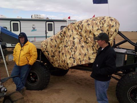 2004 Jeep Wrangler - Two men stand next to a custom off-road vehicle covered in a camouflage tarp, parked in a sandy area with a trailer in the background.