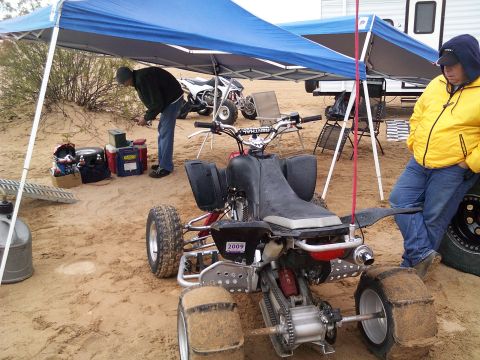 2004 Jeep Wrangler - A black four-wheel ATV is parked on the sand under a blue canopy. A person in a yellow jacket stands next to the ATV.