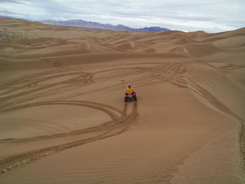 2004 Jeep Wrangler - A person rides an ATV with a red flag on a sand dune, leaving tire tracks behind them. The vast desert landscape stretches out under a clou…