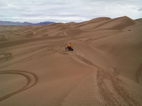 2004 Jeep Wrangler - A person in a yellow shirt rides an ATV with a red flag through a vast desert landscape with rolling sand dunes and tire tracks.