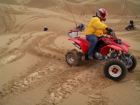 2004 Jeep Wrangler - A person in a yellow jacket and red helmet rides a red Honda ATV through sandy dunes, kicking up dust. Another ATV is visible in the backgr…