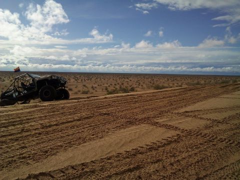 2004 Jeep Wrangler - A dark-colored off-road buggy with a roll cage is parked on a sandy dune under a partly cloudy sky. Tire tracks are visible in the foregrou…