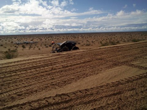 2004 Jeep Wrangler - A black off-road buggy with a flag is parked on a sand dune in a desert landscape. In the distance, several RVs and vehicles are visible.