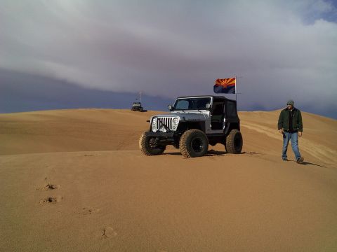 2004 Jeep Wrangler - A silver Jeep Wrangler with an Arizona flag flies on a pole behind it, parked on a sand dune with a man walking away from it. Another Jeep…