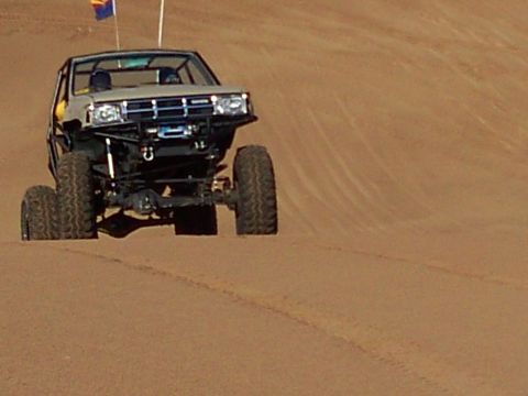2004 Jeep Wrangler - A modified off-road vehicle with large tires drives on a sandy dune. The vehicle has a roll cage and a flag mounted on the back.