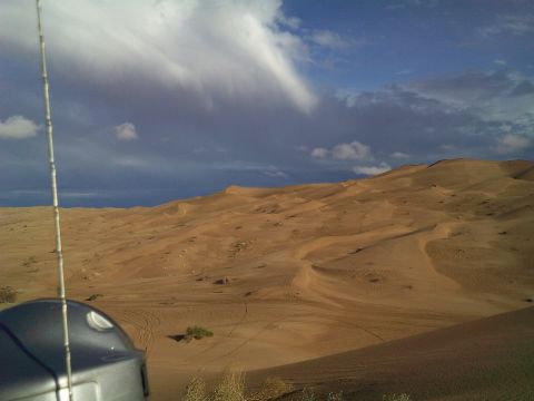 2004 Jeep Wrangler - A wide shot of sand dunes under a dramatic sky with clouds. A metallic pole and the top of a device are visible in the foreground.