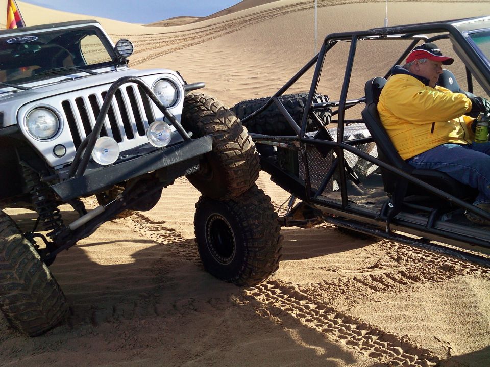 Silver Jeep Wrangler next to a dune buggy with a driver in a yellow jacket on sand dunes.
