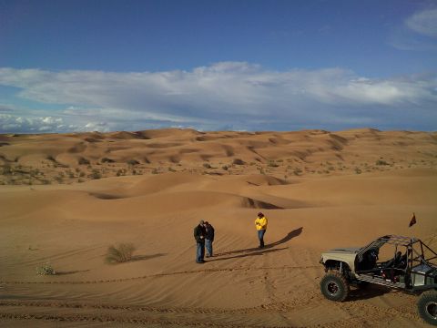 2004 Jeep Wrangler - A custom off-road buggy with a roll cage sits on a sandy dune under a blue sky with scattered clouds. Two people stand in the sand in the m…
