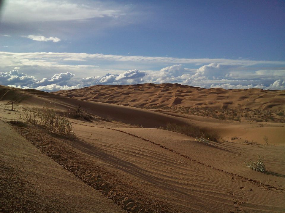 Desert sand dunes with rolling hills and a partly cloudy sky.