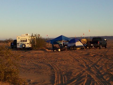 2004 Jeep Wrangler - A campsite is set up in a sandy desert landscape with multiple vehicles including a pickup truck towing a trailer with a tent and a small c…