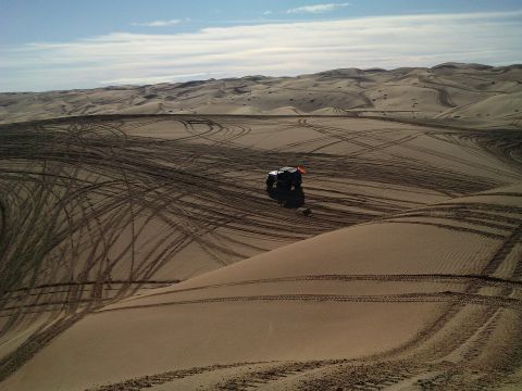 2004 Jeep Wrangler - A dark-colored off-road vehicle with an orange flag is parked on a sand dune, surrounded by numerous tire tracks from previous travel acros…