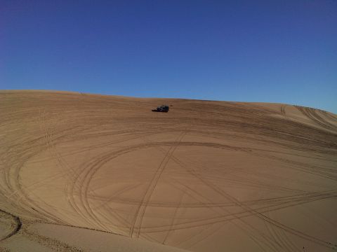 2004 Jeep Wrangler - A dark-colored Jeep Wrangler is parked on a sandy dune, leaving tire tracks across the landscape under a clear blue sky.