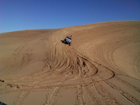 2004 Jeep Wrangler - A black Jeep Wrangler is driving down a sandy dune, leaving tire tracks in its wake under a clear blue sky.