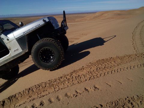 2004 Jeep Wrangler - A silver Jeep Wrangler Rubicon is shown driving on a sand dune with tire tracks visible in the sand. The vehicle is angled upwards on the d…