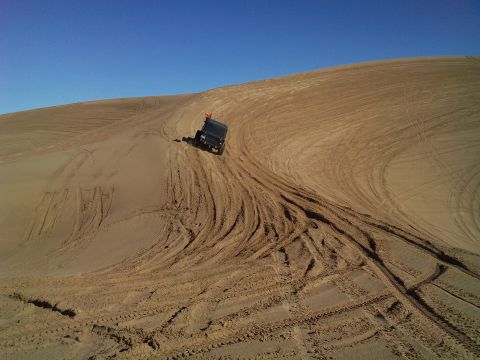 2004 Jeep Wrangler - A black Jeep Wrangler is driving down a sandy dune, leaving tire tracks in its wake under a clear blue sky.