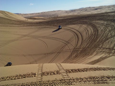 2004 Jeep Wrangler - A silver Jeep Wrangler is seen driving on a sand dune, leaving tire tracks behind it. Another smaller off-road vehicle is visible in the fo…