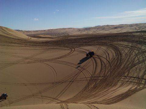 2004 Jeep Wrangler - A dark-colored ATV with an orange flag on top is shown kicking up dust on a sandy dune, leaving tire tracks behind it. Another ATV is visib…